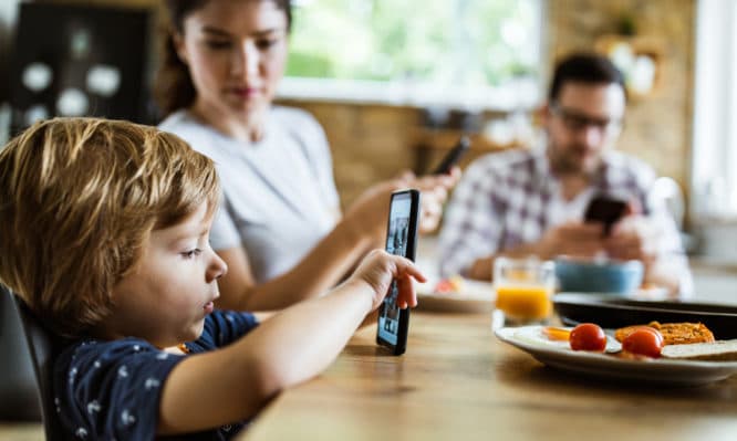 Profile view of small boy using smart phone at dining table while his parents are in the background.