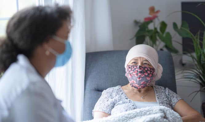 In a hospital room, an asian female cancer patient is chatting with her doctor. Both of them are wearing a mask during the coronavirus pandemic to help prevent the transfer of germs. The doctor is a female and of African American ethnicity.