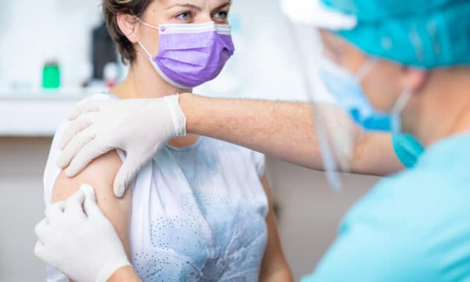 Female patient with protective face mask waiting for vaccination, doctor in surgical gloves disinfecting her arm